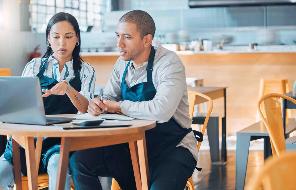 Two restaurant employees are using a laptop together to discuss business strategy at a cafe table