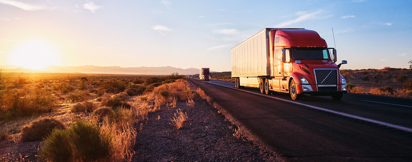 A panoramic view of a semi-truck driving on a highway through a vast desert during a sunset