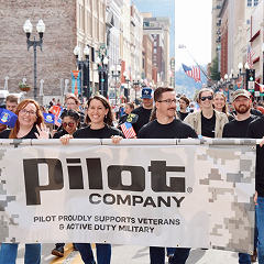 A group of smiling Pilot Company employees is marching in a parade with a banner supporting military veterans.