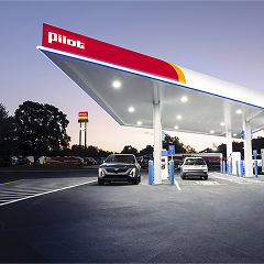 A wide-angle evening shot of a modern Pilot gas station canopy lit up with bright LED lights, featuring electric vehicle charging stations and cars parked under the structure