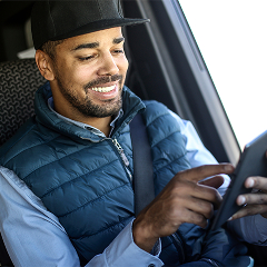 A smiling man with a beard and a black baseball cap sits in the driver's seat of a vehicle, wearing a blue quilted vest