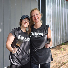 Two women in black Pilot Company t-shirts smile and pose for a photo while holding paintbrushes, likely during a community service or volunteer event