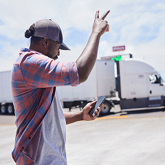 A man in a plaid shirt and baseball cap stands in a parking lot, holding a smartphone in one hand and flashing a peace sign toward a white semi-truck in the background