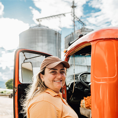 A smiling woman in a tan baseball cap and orange polo shirt stands in front of a bright orange truck