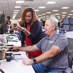 Two corporate employees are collaborating at a desk with multiple computer screens in a large, busy office.