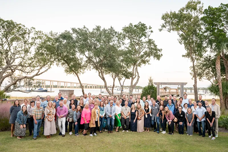 Large group of people posing outdoors near trees and water
