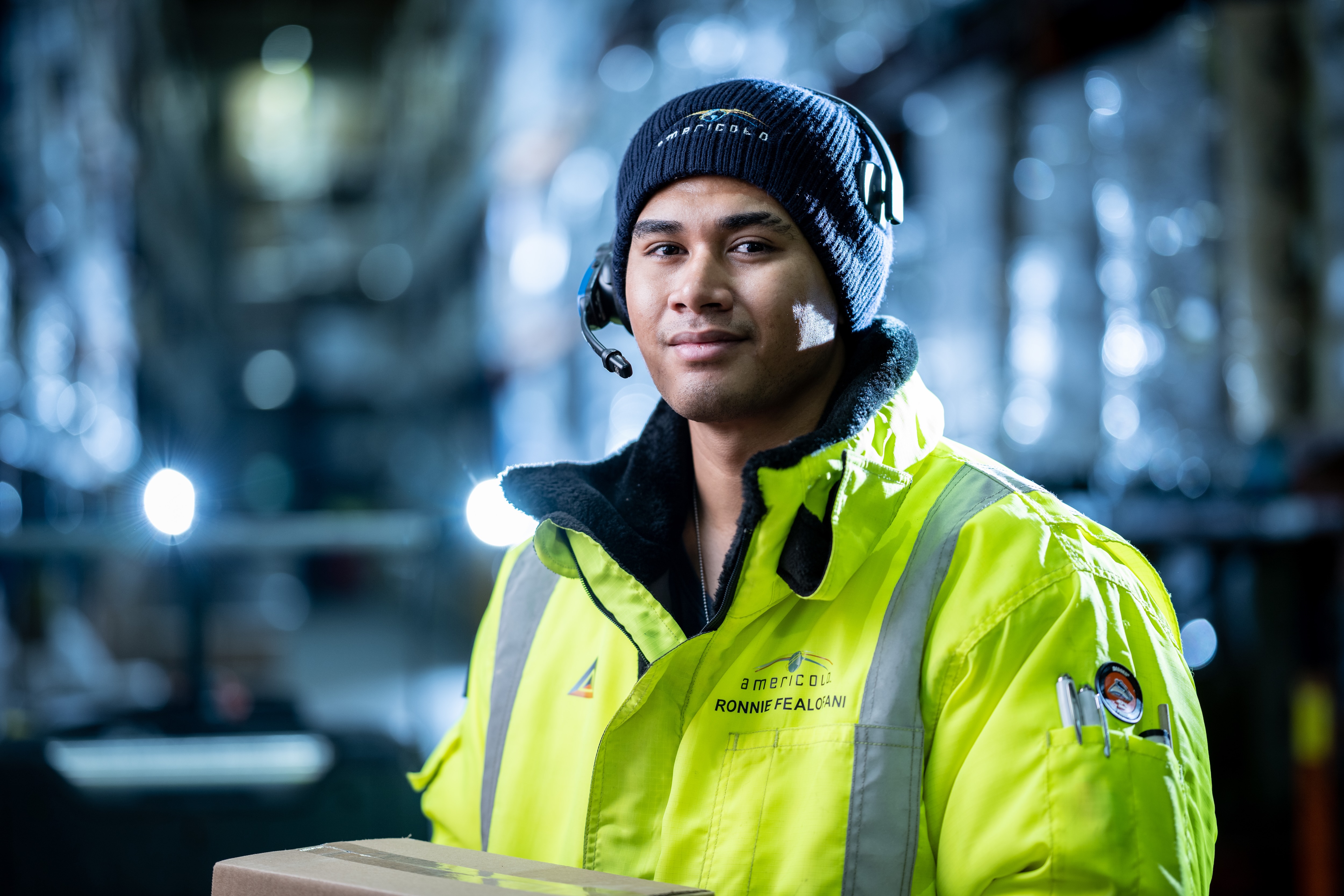 One man in yellow safety gear walk through an industrial setting, holding a box.