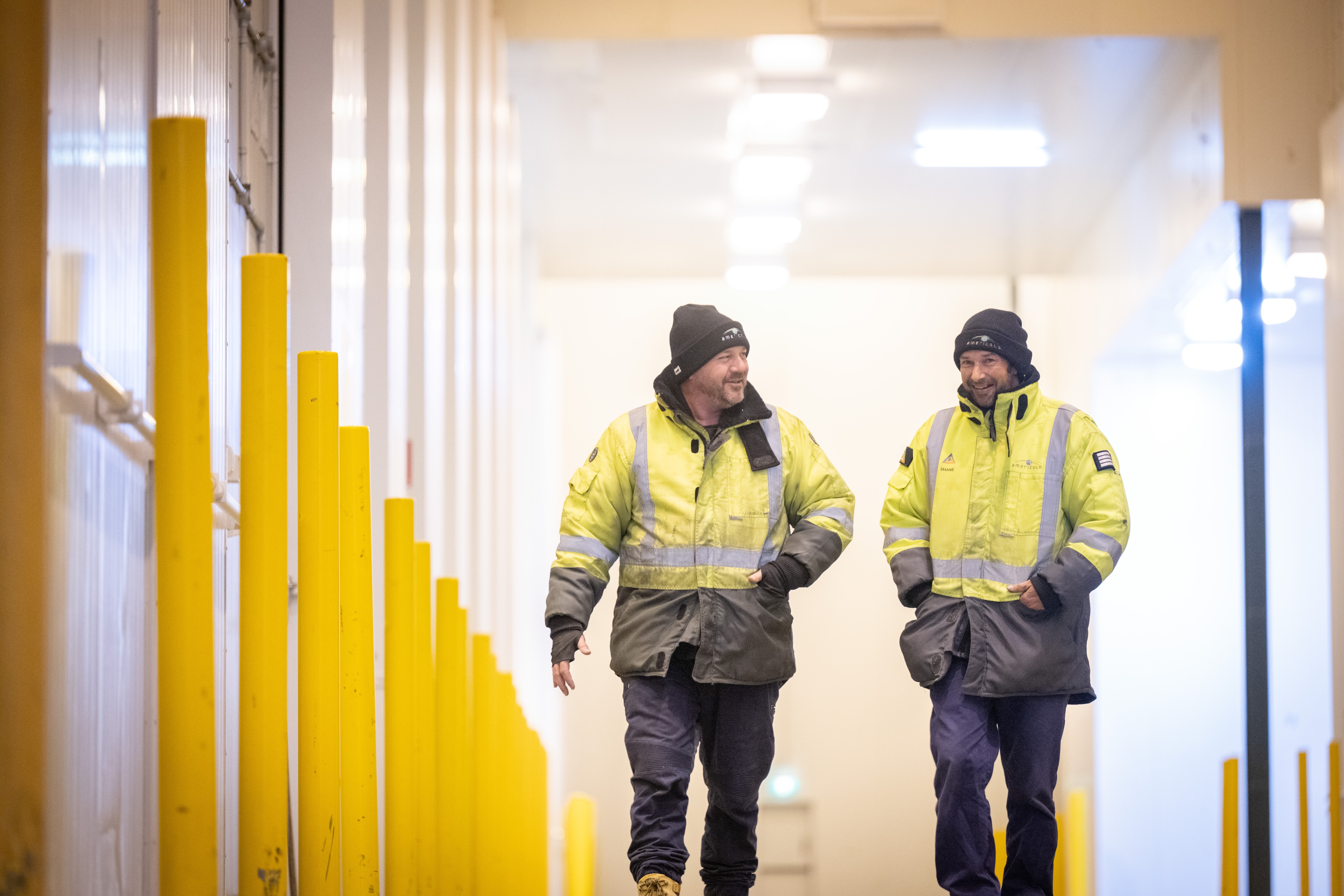 Two workers in yellow jackets walk through a hallway with yellow barriers.
