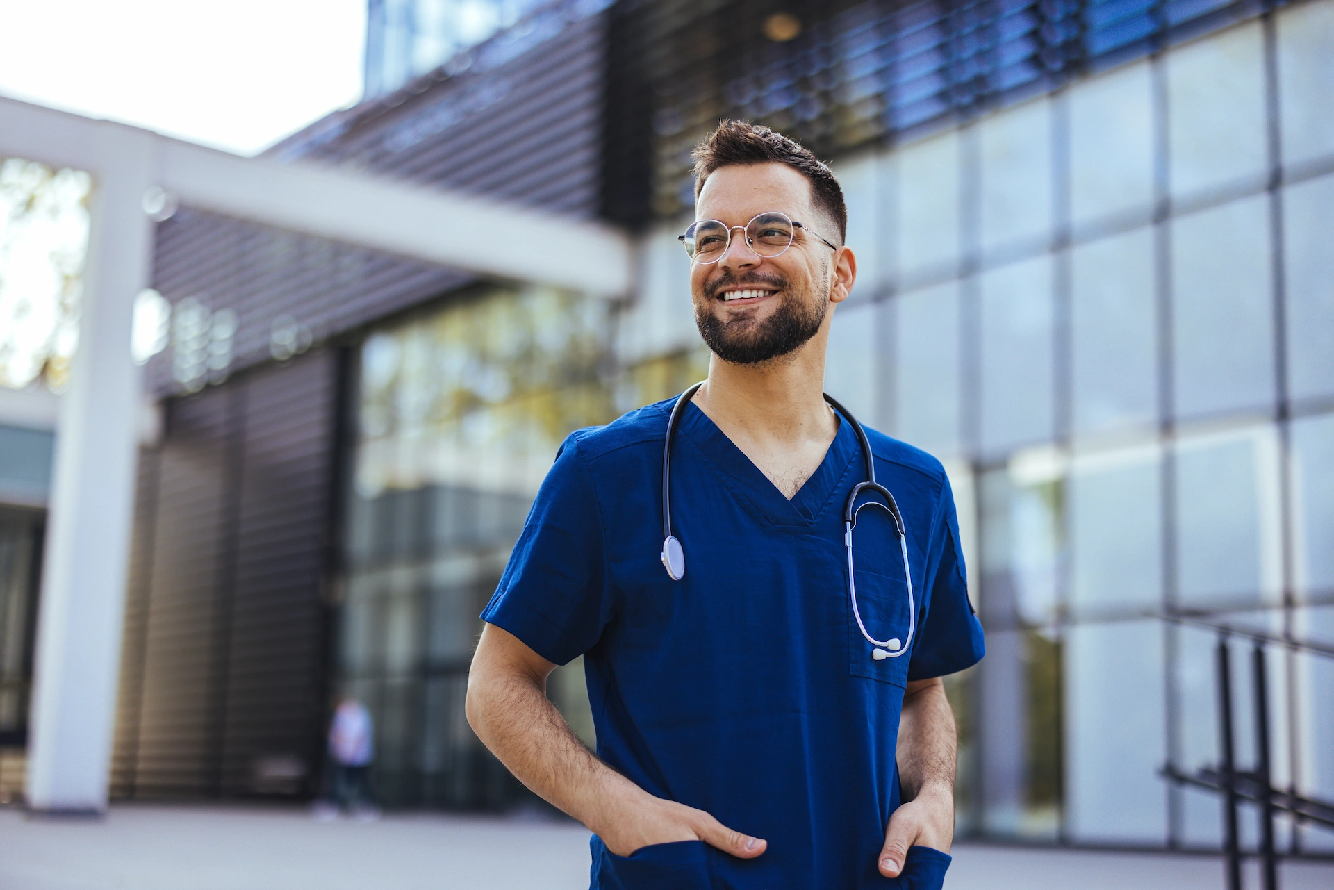 Smiling male doctor in blue scrubs with a stethoscope, beard, and glasses standing outside a modern building.